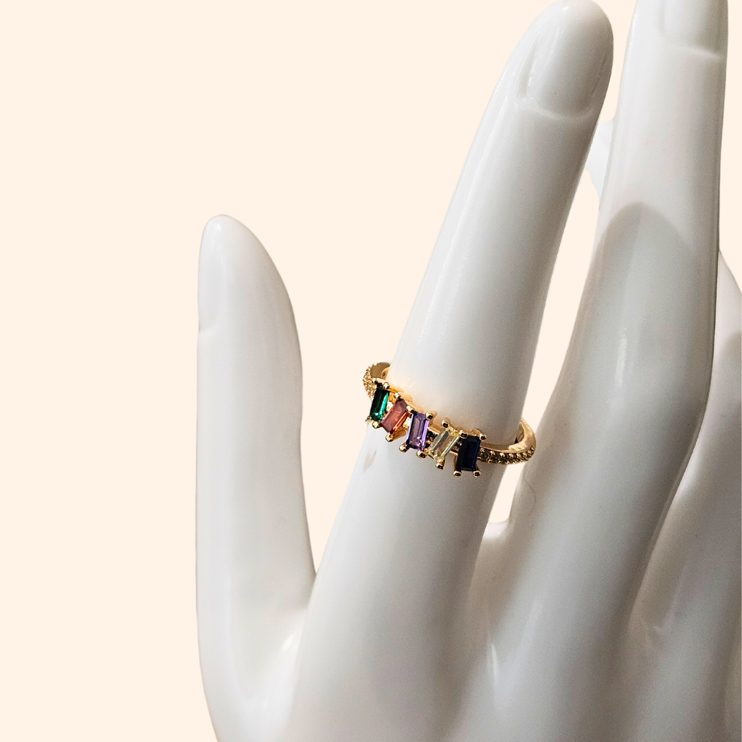 The Five Gems Gold Ring by Roots Jewellery features a dazzling arrangement of five rectangular zircons in vibrant hues—red, green, purple, blue, and clear—set diagonally.