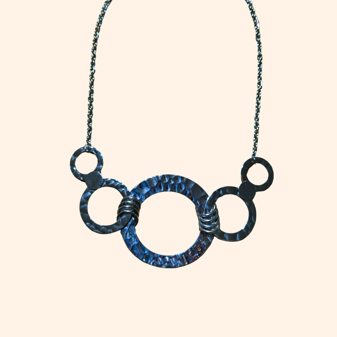 A person wearing a denim jacket and a ribbed white shirt is accessorized with the Roots Jewellery Golden/Silver Rings Statement Necklace Set, adding a bold geometric design to their look.