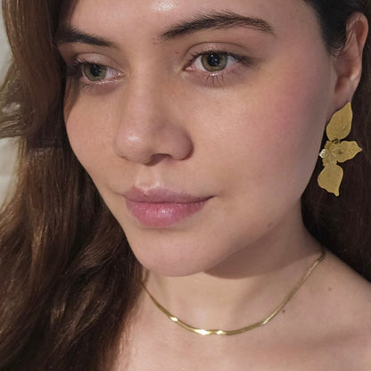 The Golden Leaf Charm Earrings by Roots Jewellery, showcasing intricate leaf designs, rest gracefully on a light beige surface and are equipped with durable stainless steel backings for enduring sophistication.