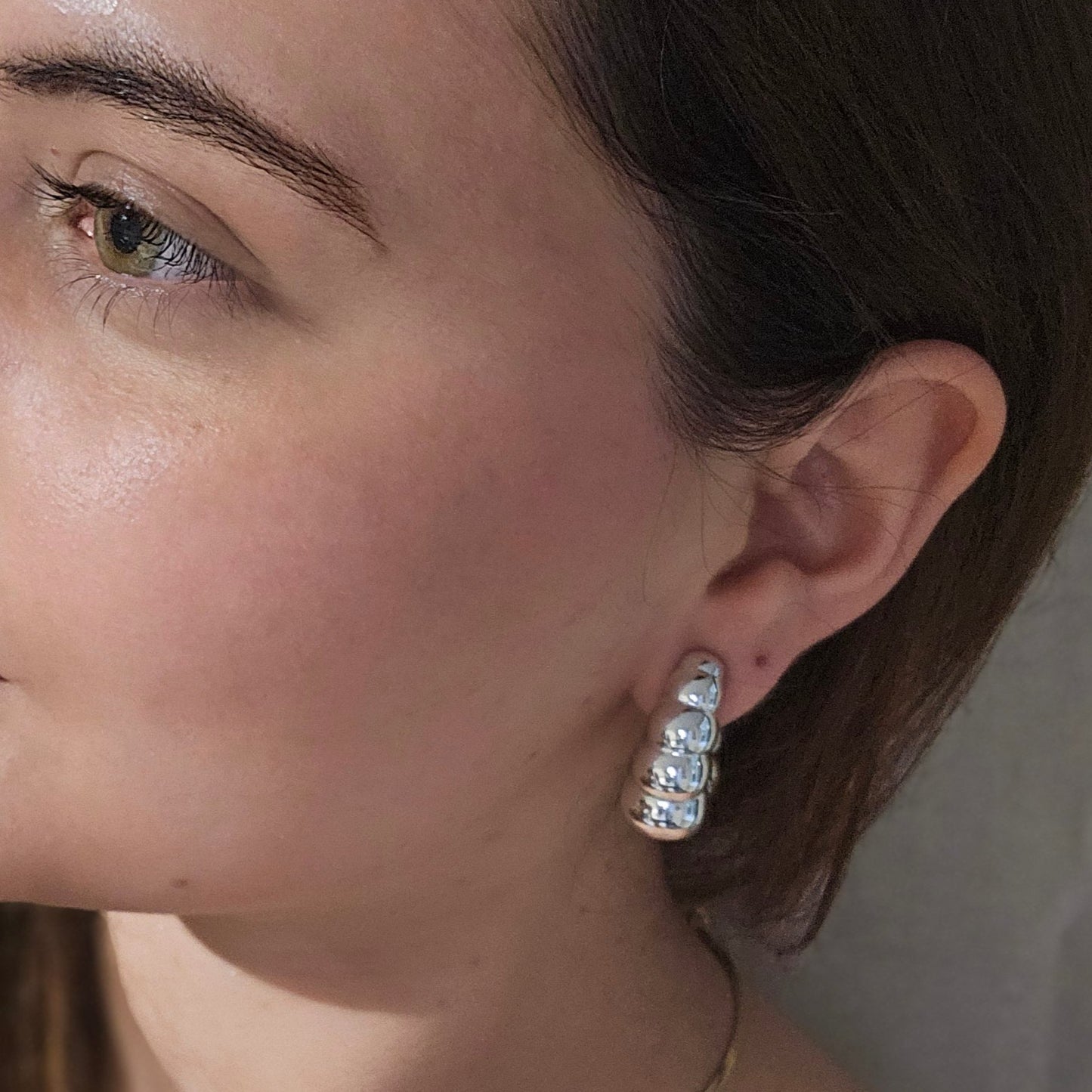 Displayed on a reflective surface, the Silver Bubble Crescent Hoop Earrings by Roots Jewellery showcase their bubble-like design. These elegant, rhodium-plated silver earrings are enhanced by a background featuring white sculptural decor and gold accents, adding an air of sophistication.