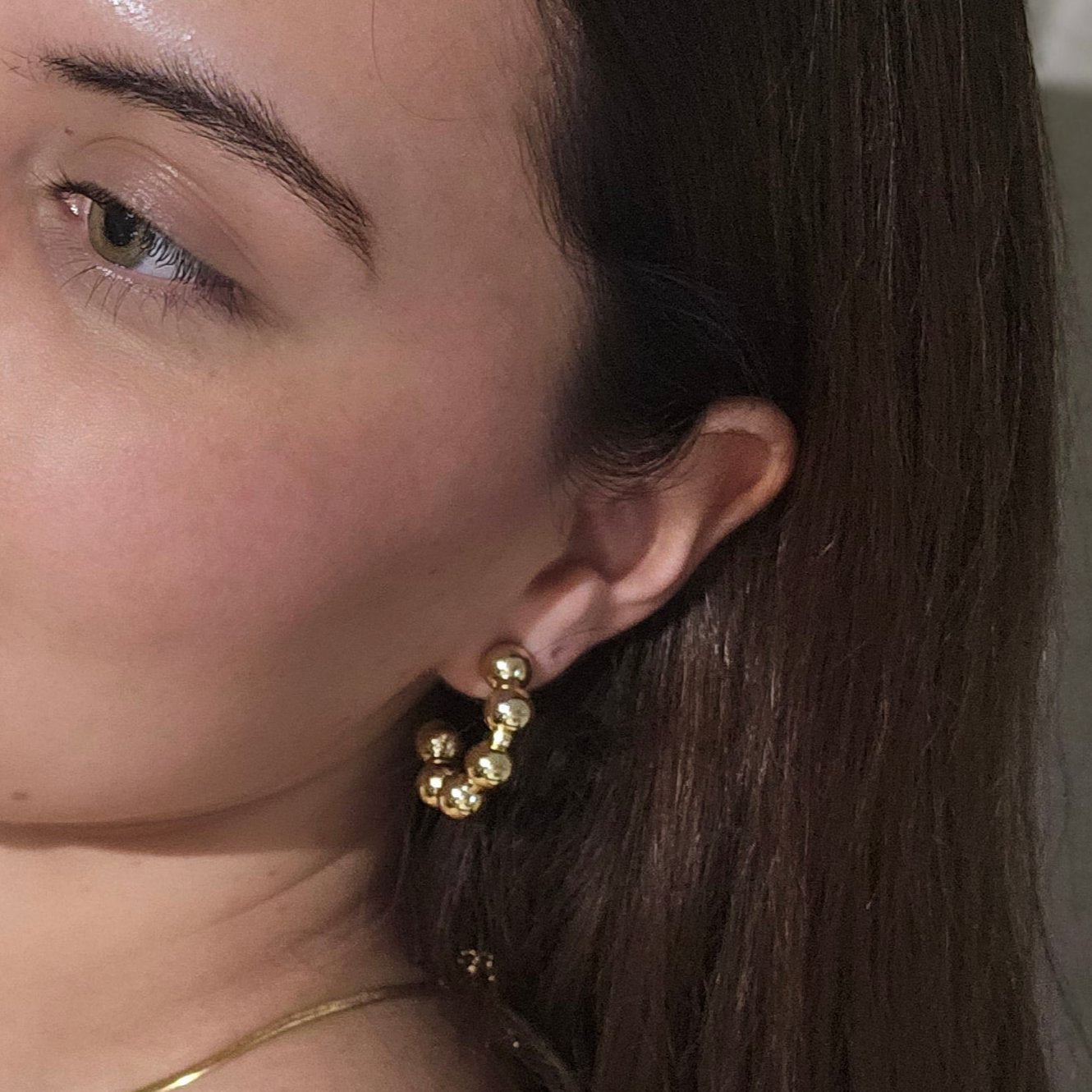 Close-up of a person's ear adorned with stylish Roots Jewellery hoops, showcasing Gold Ball Hoops Earrings. A portion of the face is visible, highlighting the jewelry's elegance with its gold and rhodium plating.