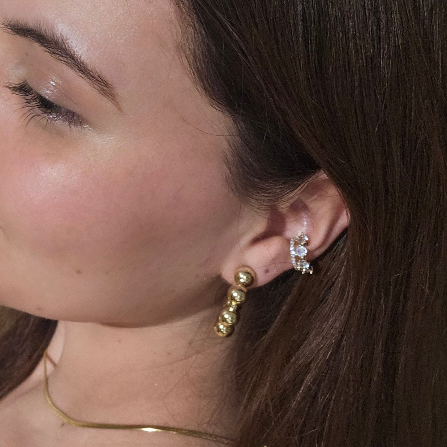 Close-up of a person's ear adorned with stylish Roots Jewellery hoops, showcasing Gold Ball Hoops Earrings. A portion of the face is visible, highlighting the jewelry's elegance with its gold and rhodium plating.