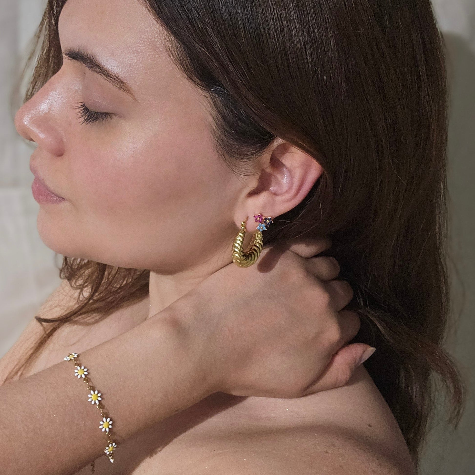 A woman with long brown hair is wearing a simple gold necklace and colorful Triple Flower Gold Earrings by Roots Jewellery, looking to the side.