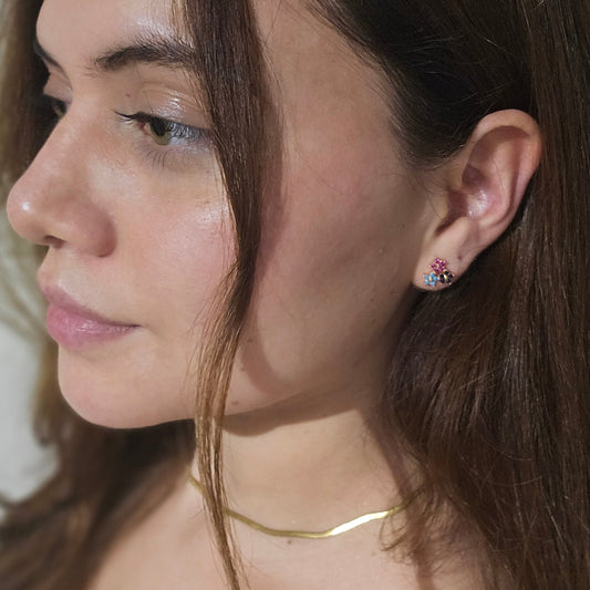 A woman with long brown hair is wearing a simple gold necklace and colorful Triple Flower Gold Earrings by Roots Jewellery, looking to the side.