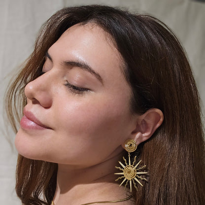 A woman with long brown hair is wearing Roots Jewellery's Sunny Gold Earrings and a necklace, looking to the side against a plain background.