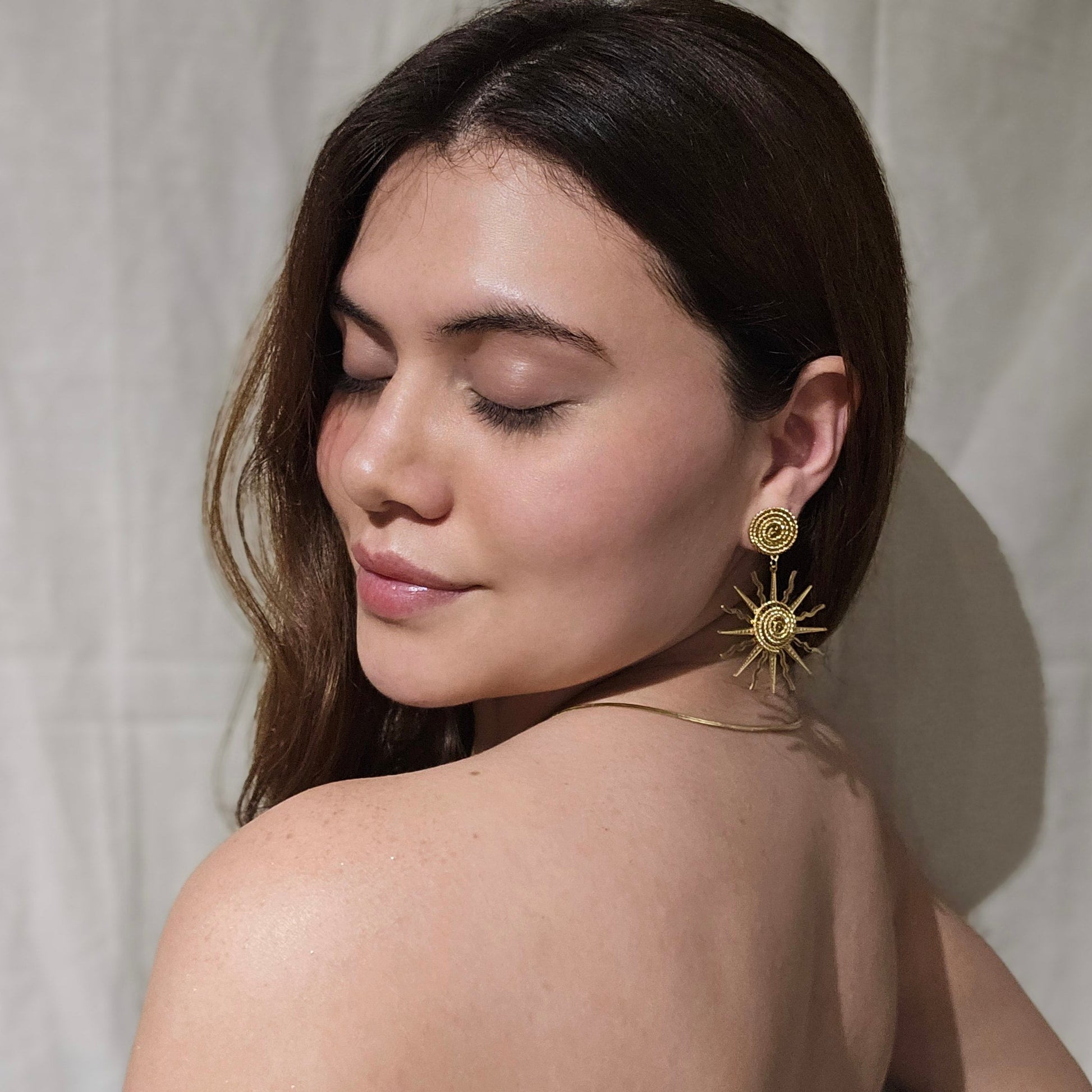 A woman with long brown hair is wearing Roots Jewellery's Sunny Gold Earrings and a necklace, looking to the side against a plain background.