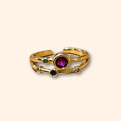 Discover the Infinite Gold Ring by Roots Jewellery, a breathtaking gold creation adorned with a prominent central purple gemstone and four smaller multicolored stones on an adjustable split band. Enhanced with rhodium plating for enduring brilliance, it is elegantly showcased against a beige backdrop.