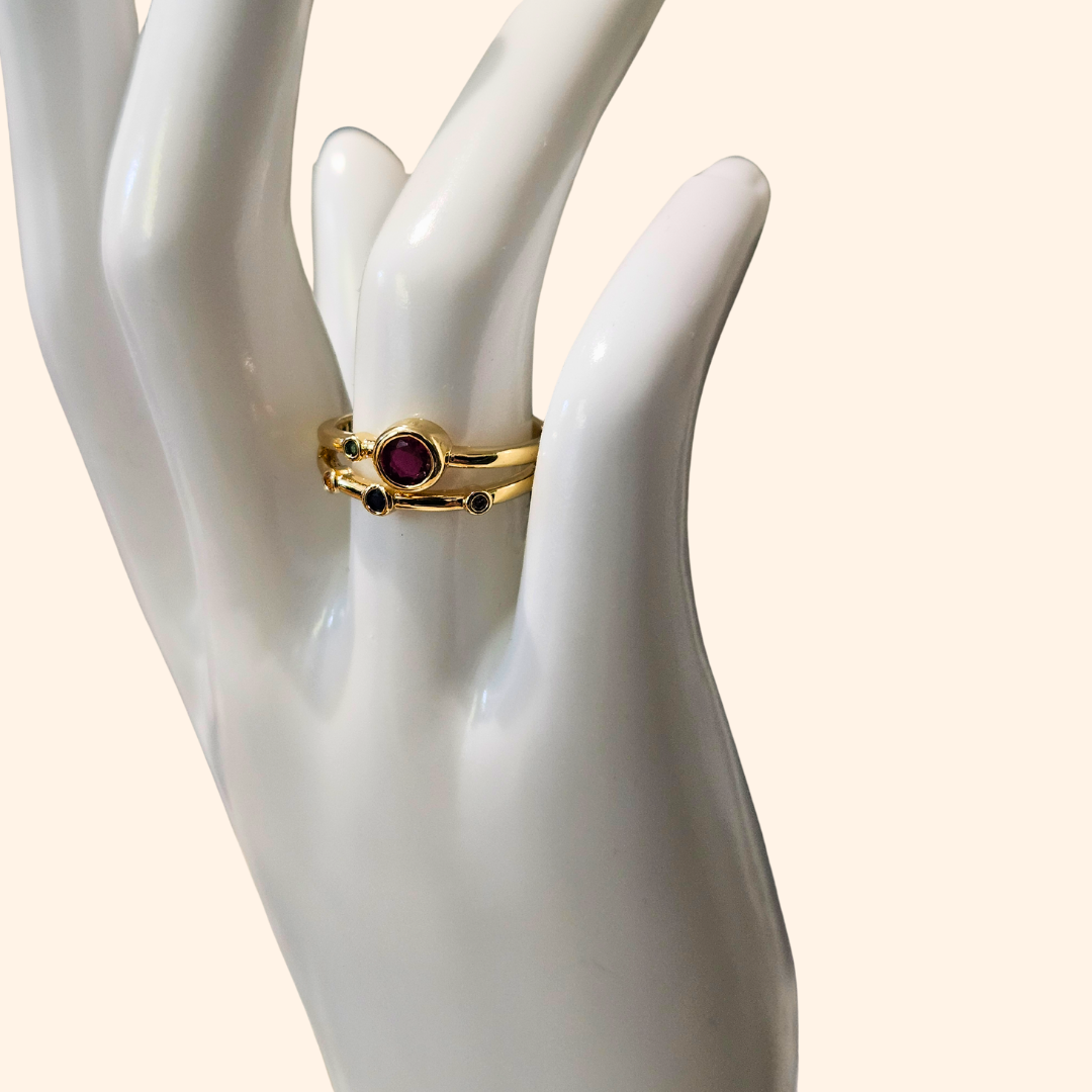 Discover the Infinite Gold Ring by Roots Jewellery, a breathtaking gold creation adorned with a prominent central purple gemstone and four smaller multicolored stones on an adjustable split band. Enhanced with rhodium plating for enduring brilliance, it is elegantly showcased against a beige backdrop.