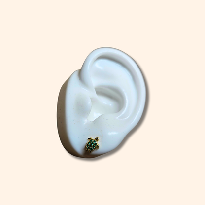 The Zirconia Turtle Gold Earrings by Roots Jewellery, featuring a trio of small turtles embellished with green, black, and pink zircons, are elegantly showcased on a white ear-shaped stand.