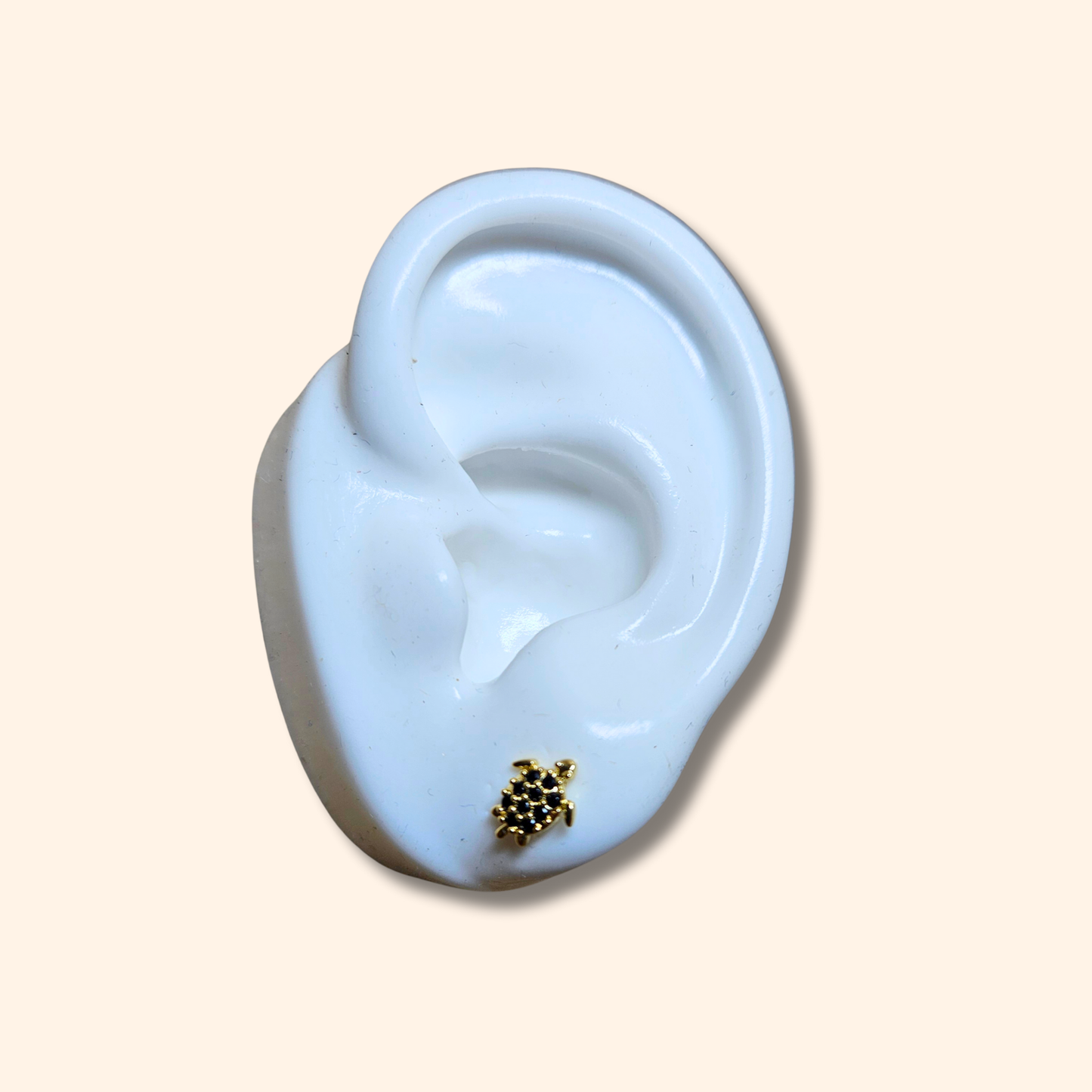 The Zirconia Turtle Gold Earrings by Roots Jewellery, featuring a trio of small turtles embellished with green, black, and pink zircons, are elegantly showcased on a white ear-shaped stand.