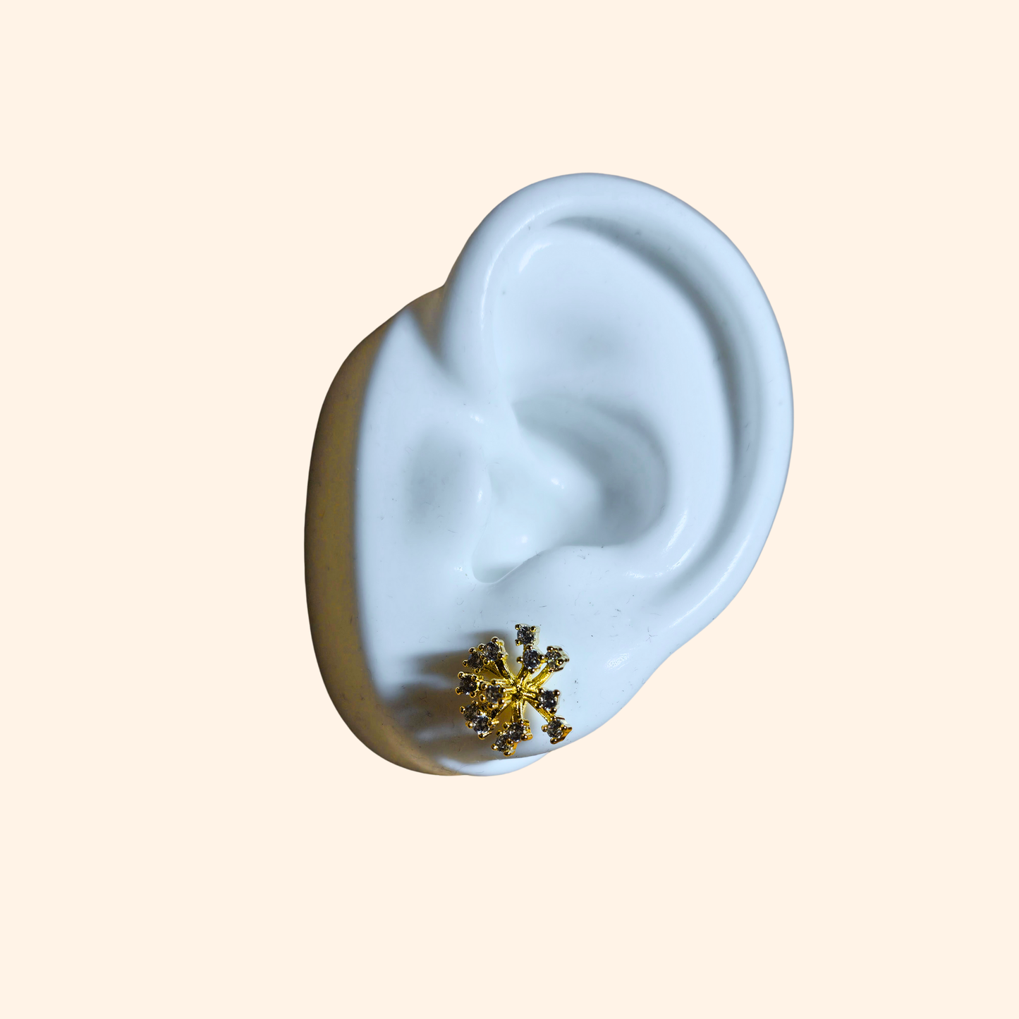 A white ceramic ear presents the Golden Snowflake Stud Earrings by Roots Jewellery, adorned with stainless steel gold plating and shimmering zircons, set against a beige background.