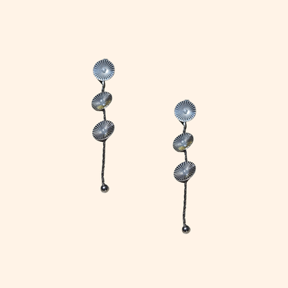 The Cosmic Earrings by Roots Jewellery feature silver and gold drop designs with circular patterns and elongated elements, providing a contemporary appeal inspired by cosmic motifs. They are showcased against a neutral backdrop.