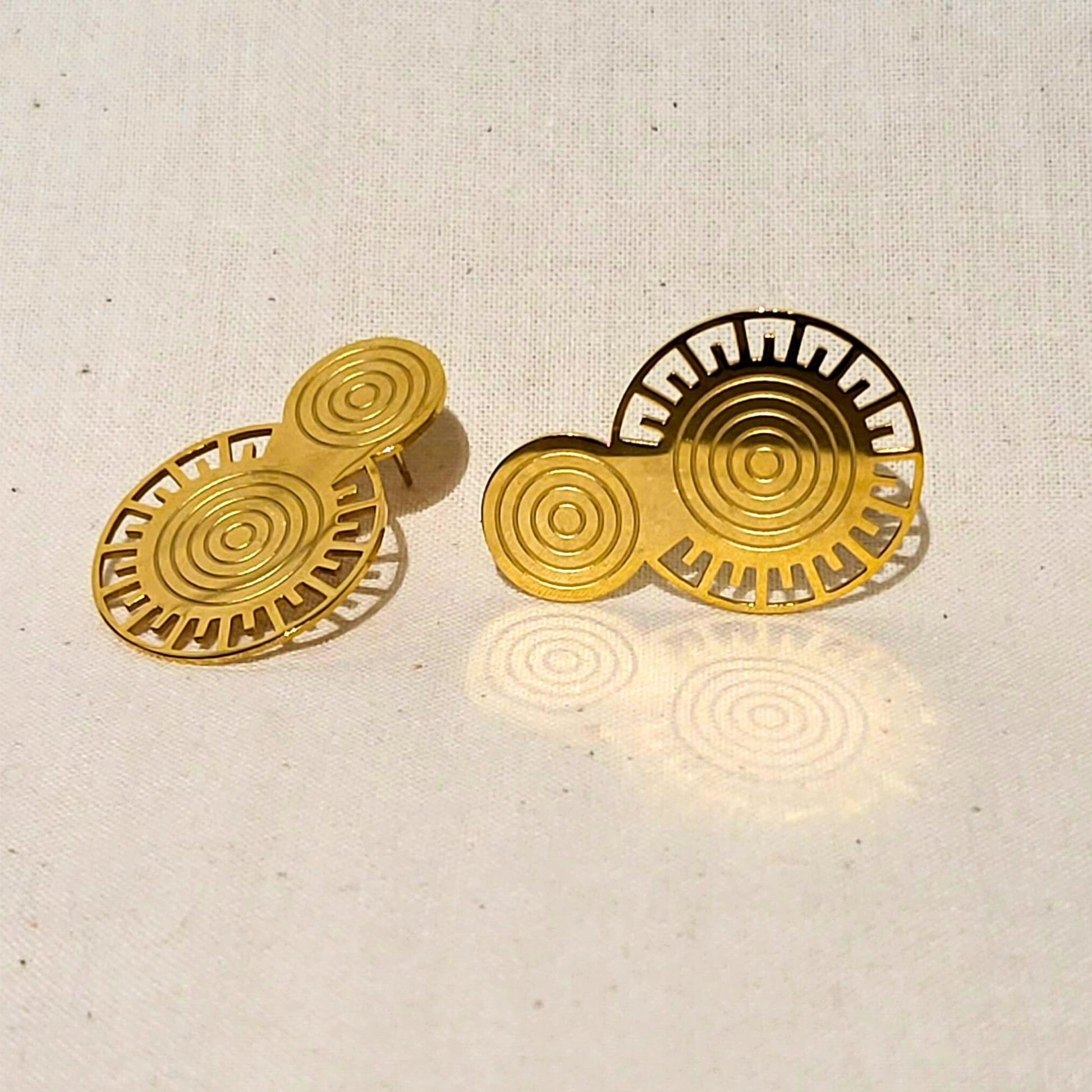 The Golden Orbit Earrings by Roots Jewellery feature two gold, circular objects adorned with abstract patterns on a beige background, embodying a modern design. Their metallic gold finish adds an elegant and stylish allure.