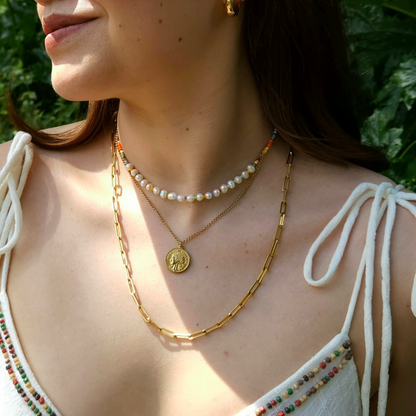 A person showcases a striking look with Roots Jewellery's 3 Set Necklace with Pearls, adorned with gleaming gold chains, lustrous pearls, and a coin pendant. The attire is enhanced by string shoulder details and multicolored bead accents. Dangling earrings complete the ensemble against the lush green foliage backdrop.