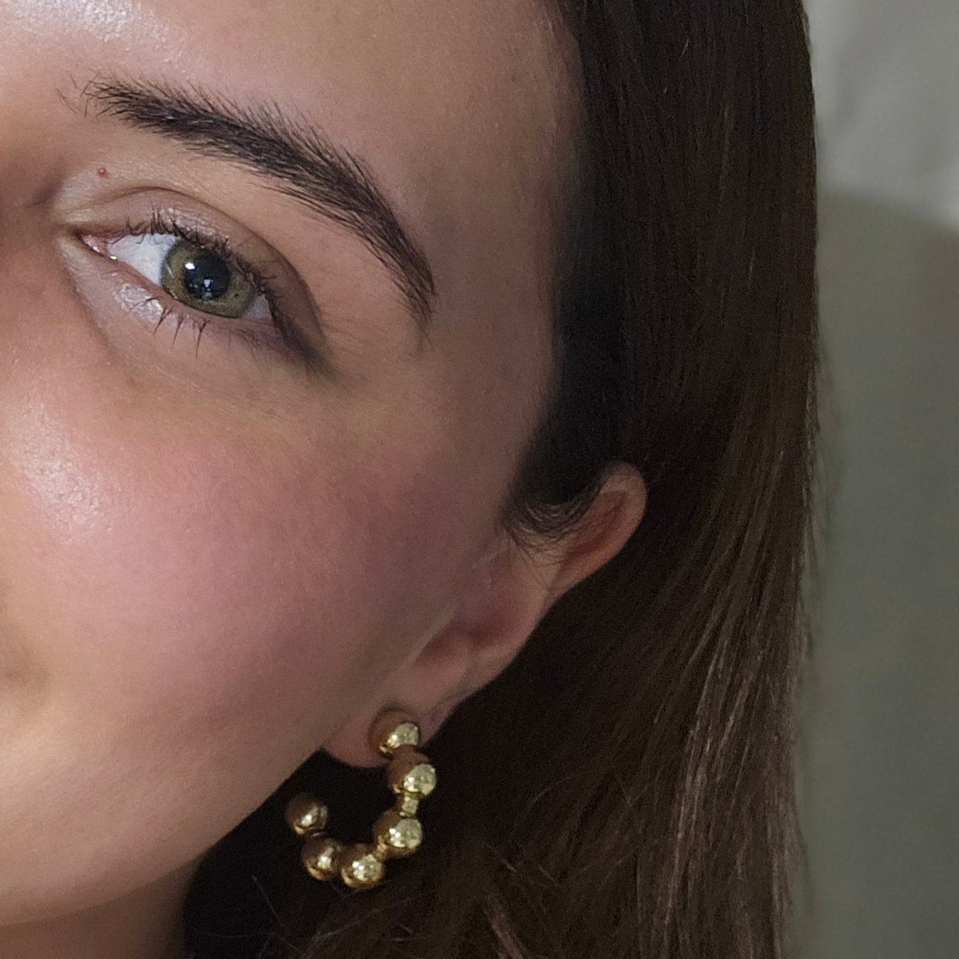 Close-up of a person's ear adorned with stylish Roots Jewellery hoops, showcasing Gold Ball Hoops Earrings. A portion of the face is visible, highlighting the jewelry's elegance with its gold and rhodium plating.