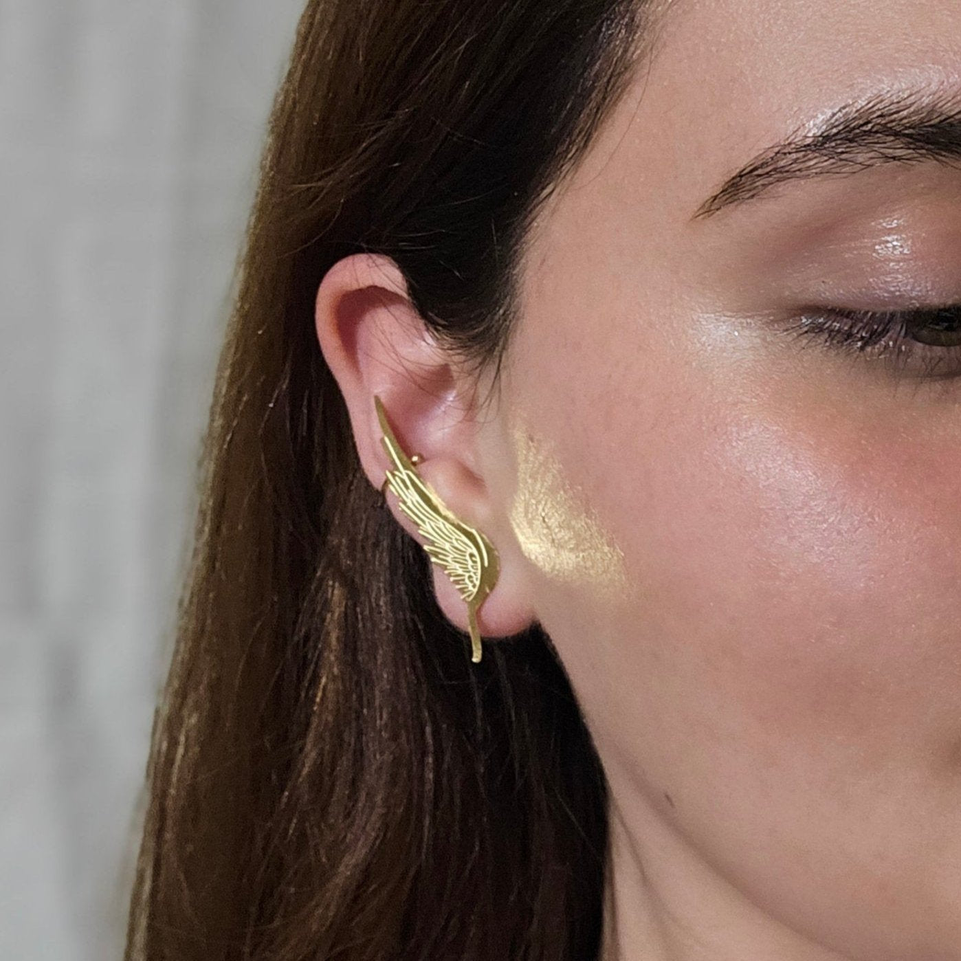 White ceramic ear model adorned with an elegant gold wing-shaped ear cuff, inspired by the Golden Angel Wings Earrings from Roots Jewellery, displayed on a beige background.