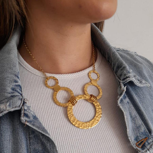 A person wearing a denim jacket and a ribbed white shirt is accessorized with the Roots Jewellery Golden/Silver Rings Statement Necklace Set, adding a bold geometric design to their look.