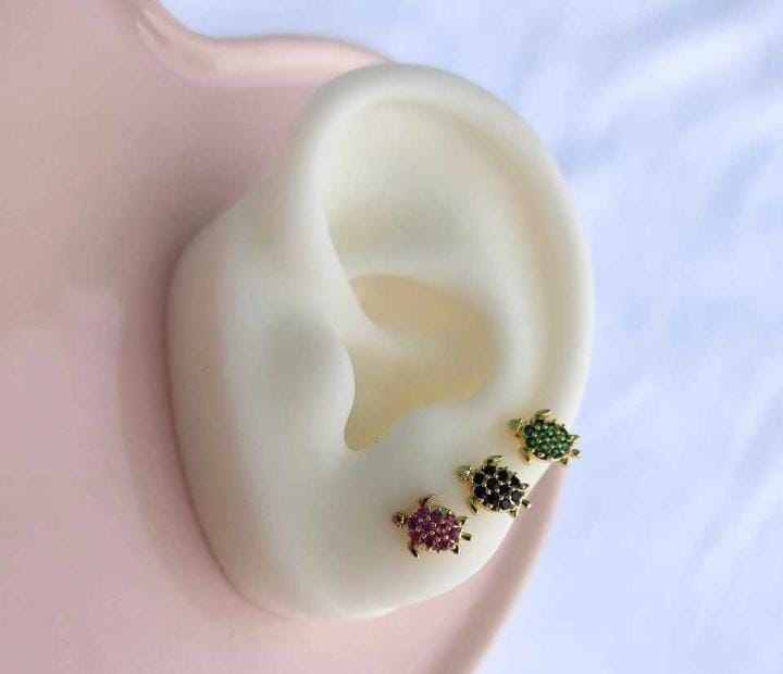 The Zirconia Turtle Gold Earrings by Roots Jewellery, featuring a trio of small turtles embellished with green, black, and pink zircons, are elegantly showcased on a white ear-shaped stand.