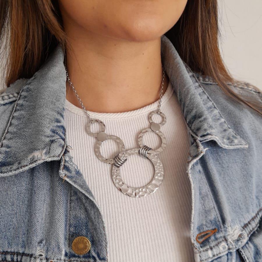 A person wearing a denim jacket and a ribbed white shirt is accessorized with the Roots Jewellery Golden/Silver Rings Statement Necklace Set, adding a bold geometric design to their look.