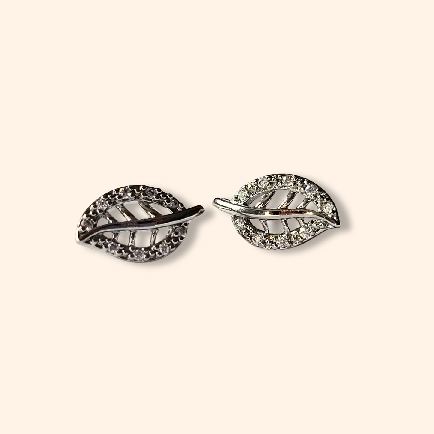 Silver Leafs Earrings by Roots Jewellery: These distinctive leaf-shaped earrings are adorned with exquisite crystal detailing and crafted from durable stainless steel, elegantly presented against a beige background.