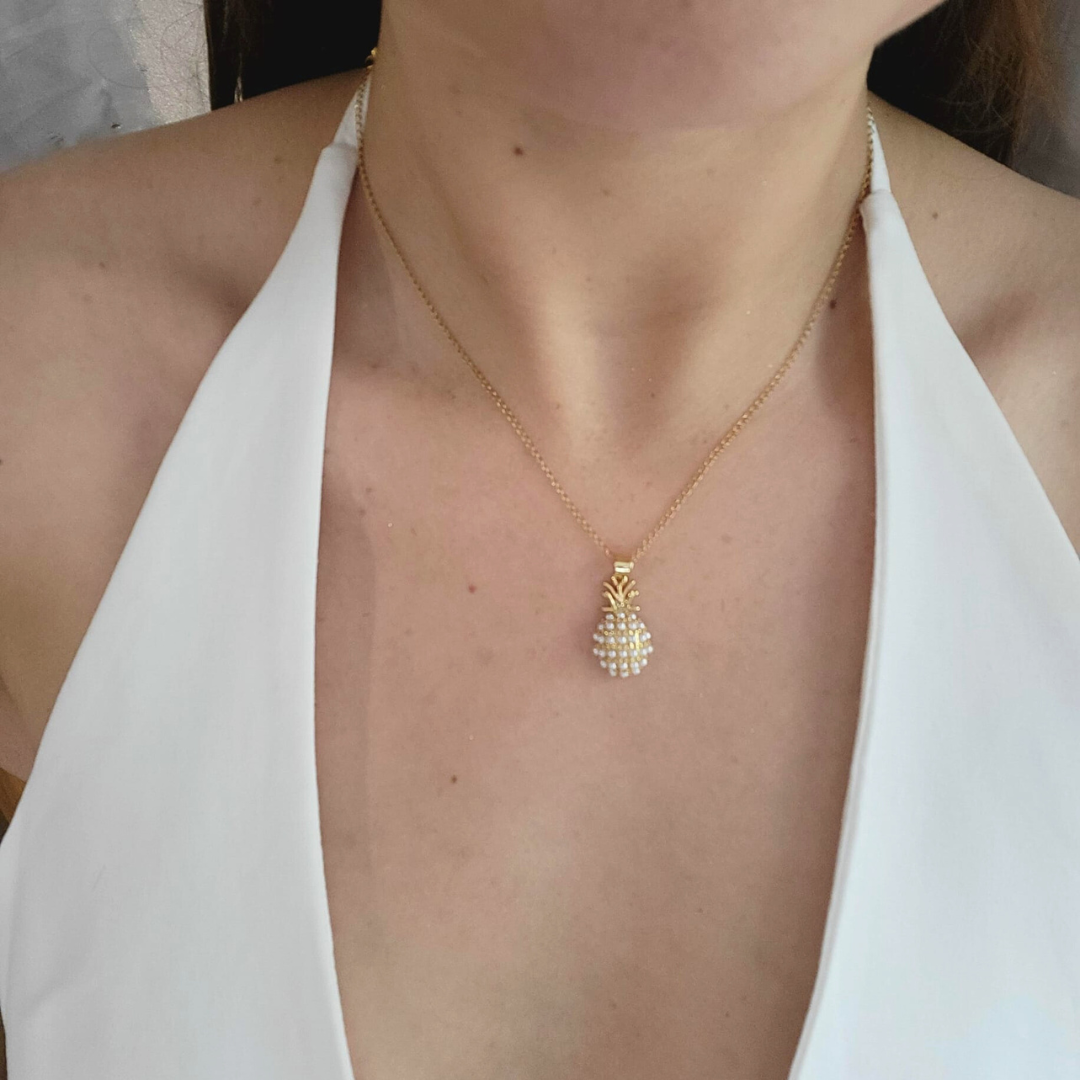 A woman dazzles in a Roots Jewellery Pineapple Necklace paired with matching earrings and a white halter top, showcasing the unique design's playful charm. The jewelry set's gold-plated finish adds a touch of elegance to the tropical theme.