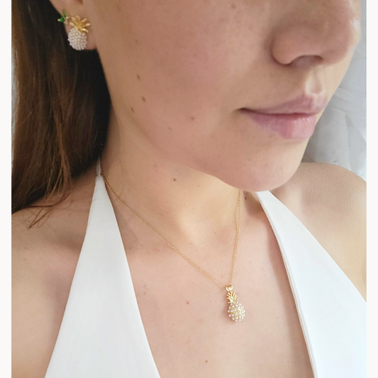 A woman dazzles in a Roots Jewellery Pineapple Necklace paired with matching earrings and a white halter top, showcasing the unique design's playful charm. The jewelry set's gold-plated finish adds a touch of elegance to the tropical theme.