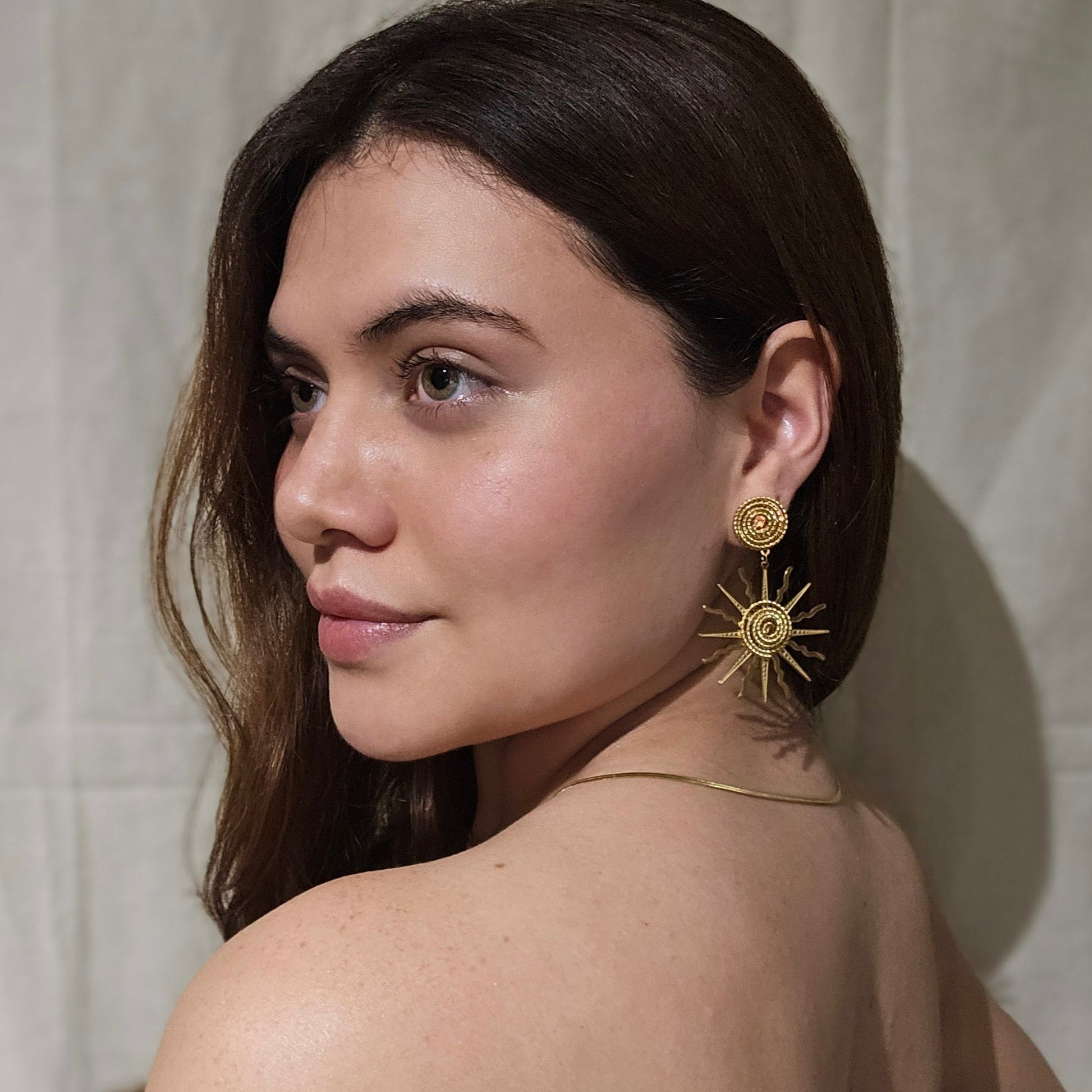 A woman with long brown hair is wearing Roots Jewellery's Sunny Gold Earrings and a necklace, looking to the side against a plain background.