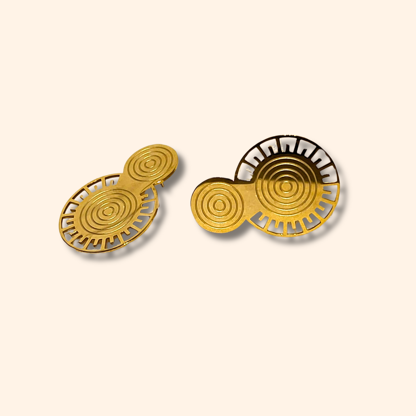 The Golden Orbit Earrings by Roots Jewellery feature two gold, circular objects adorned with abstract patterns on a beige background, embodying a modern design. Their metallic gold finish adds an elegant and stylish allure.