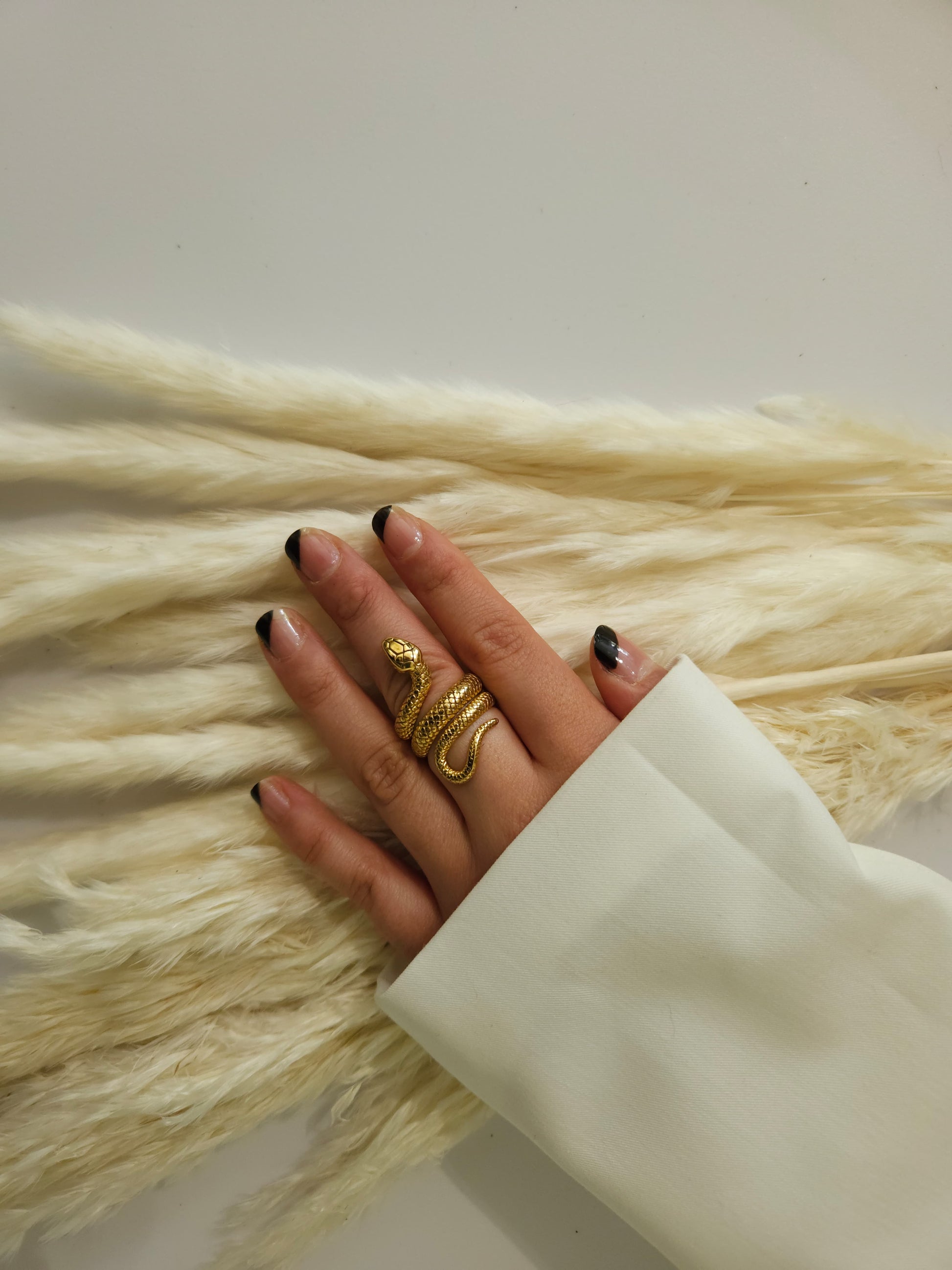 A hand adorned with a Golden Snake Ring by Roots Jewellery and black-tipped nails rests gracefully on soft, white pampas grass.